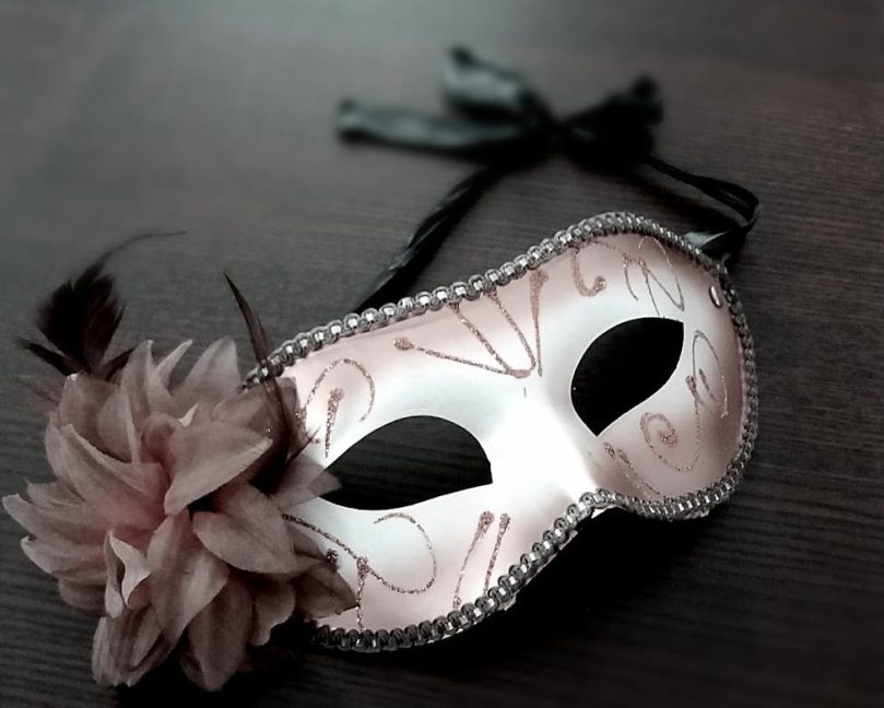 A decorative masquerade mask with a pink flower adornment, placed on a dark wooden surface.